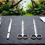 A premium 5-piece stainless steel aquascaping tool kit laid out on a dark slate surface in front of a lush planted aquarium.
